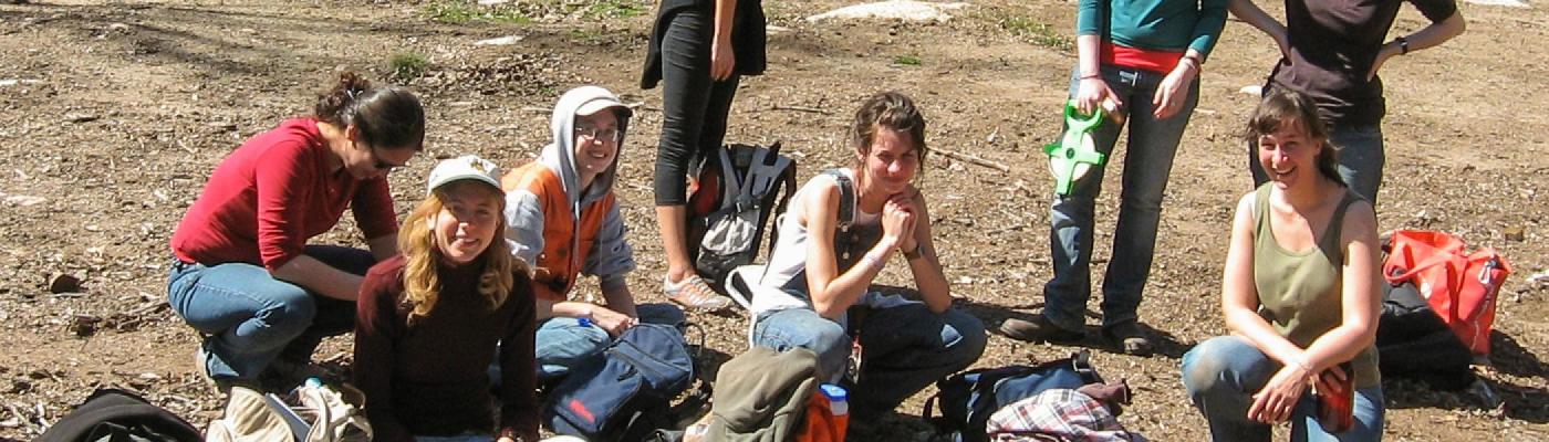 A group of students sitting outside in a wooded area on the ground in, smiling at the camera