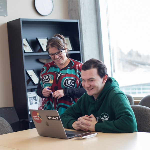 A student sitting and an instructor standing working together on a computer smiling