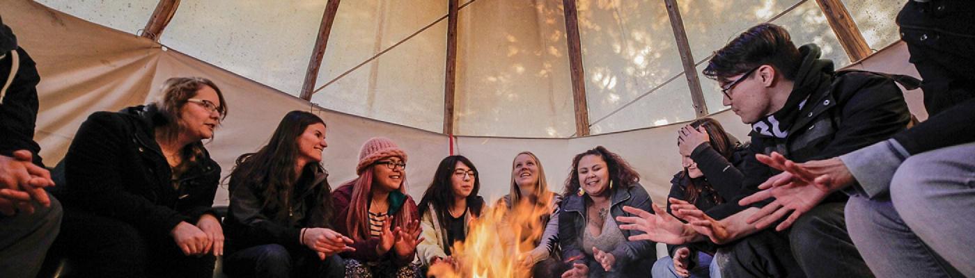 A group of students sitting around a fire in a tipi listening to a professor speak