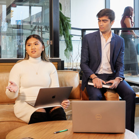 Two students sitting on a couch while speaking with colleauges in a shared working space