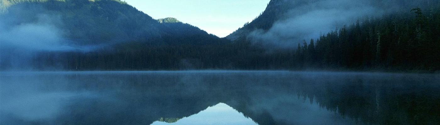 Picture taken from the side of a lake, with forest and mountains in the distance.