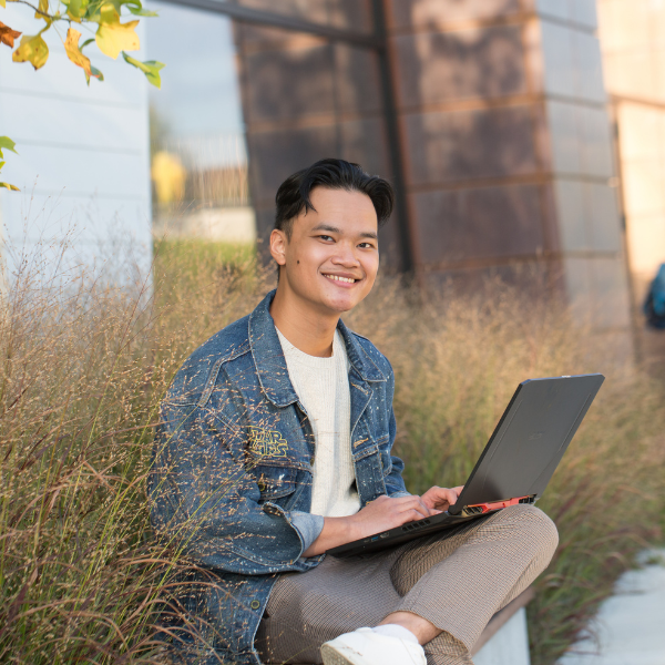Student sits outside next to a long grass garden on his laptop