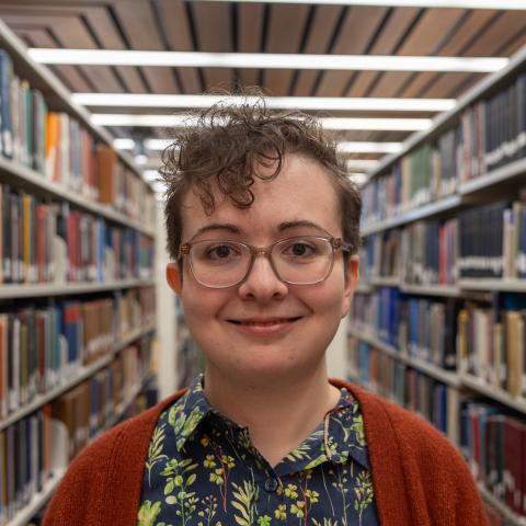 A person with short curly hair, glasses, and a floral button-up shirt paired with an orange sweater, poses with a gentle smile in a library