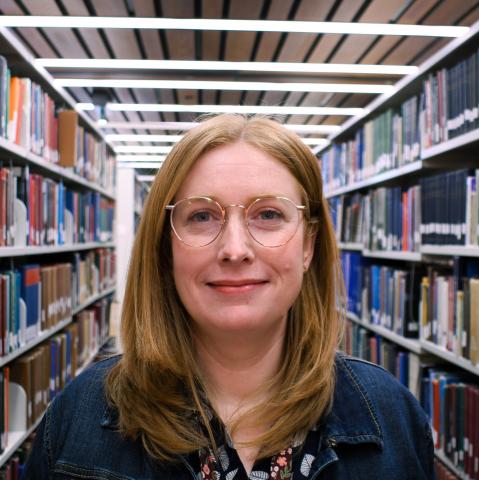 A woman with shoulder-length red hair and round glasses, wearing a denim jacket, smiles softly while standing in a library.