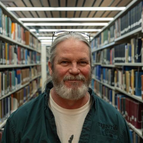 An man with long gray hair and a beard, wearing glasses and a green jacket, poses with a slight smile in a library setting.