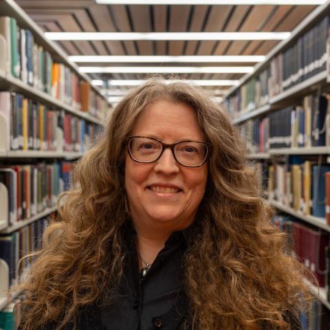 A woman with long, curly gray hair and glasses, wearing a black shirt, smiles as she stands in a library aisle.