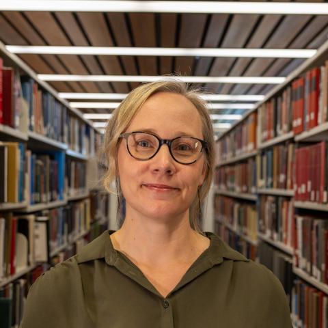 A woman with blonde hair and glasses, wearing a green shirt, smiles softly while standing in a library aisle.