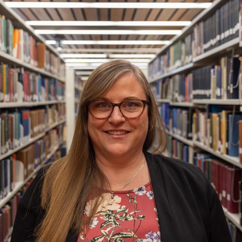 A woman with long blonde hair, wearing glasses and a floral blouse, smiles warmly in a library aisle.