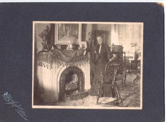 Matted photograph of a woman standing by a fireplace and a spinning wheel in what looks to be the 19th century