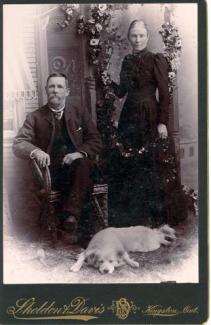 Black and white photograph of a man, woman and a dog, posing. At the bottom, gold letters say Sheldon & Davis, Kingston, Ontario