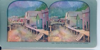Two nearly identical photographs mounted together on a board that says "1326. Street on Ocean Front, Juneau City, Alaska." The hand-coloured photographs are of houses on stilts with mountains in the background.