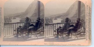 Two nearly identical black and white photographs mounted on teh same board. Image is of two men sitting in chairs on a balcony looking at a landscape with water, trees, and a mountain. There is text along the bottom and sides