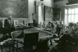 Black and white photograph of a man speaking to a group assembled in an old home, surrounded by books and art