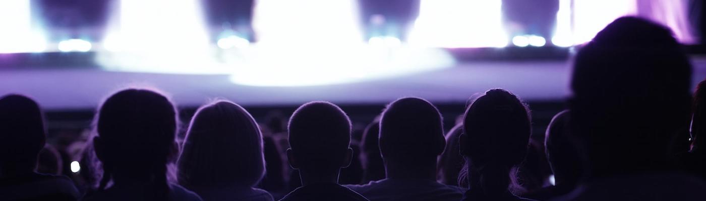 The view of a stage with bright lights from behind a row of people in a theatre.