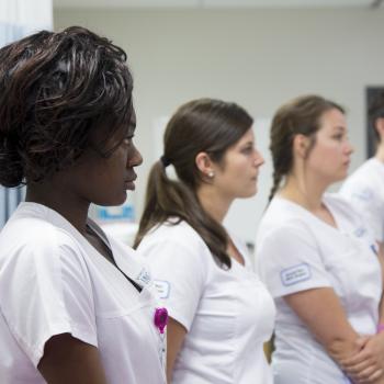 Four students standing in a line, all wearing scrube, looking at an instructor (not pictured)