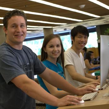 three students standing behing a desk, each with a computer monitor in-front of them, smiling at the camera