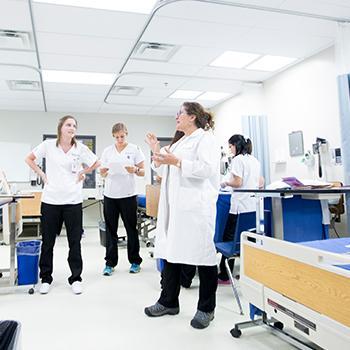 Three nursing students and one instructor in nursing lab, wearing uniforms, standing and talking.
