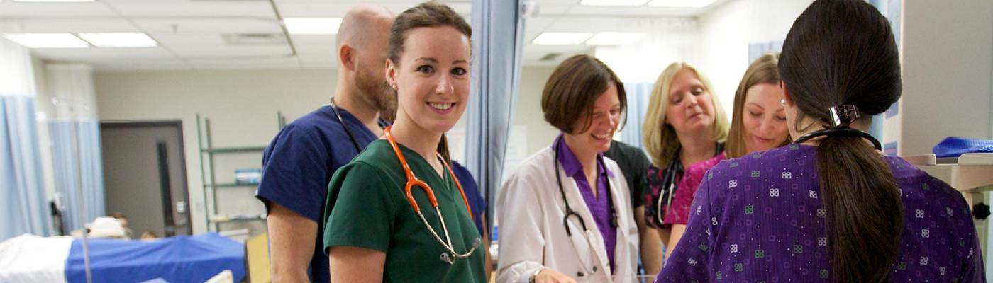 Female nursing student in scrubs smiling at camera with other students and professors in the background.