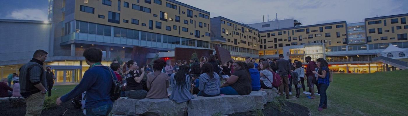 alt="Gathering of students seated in a circle facing away from camera with Peter Gzowski building in the background"