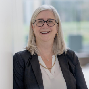 Photograph of Dr. Tana Allen smiling. Tana is wearing a suit jacket and glasses. The background is bright.