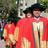 A long line of students in red convocation gowns waling along the Lady Eaton College brdige, smiling at the camera in the morning sun