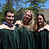 Students sitting in chairs outside on the podium wearing green gowns, smiling at the camera