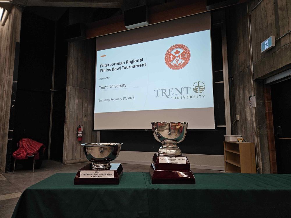 The Peterborough Regional Ethics Bowl tropheys sit in front of a screen that advertises the event.
