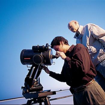 A student looking into a telescope with a professor behind him looking at the sudent