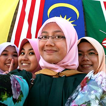 Young women wearing hijab in convocation robes in front of multiple flags