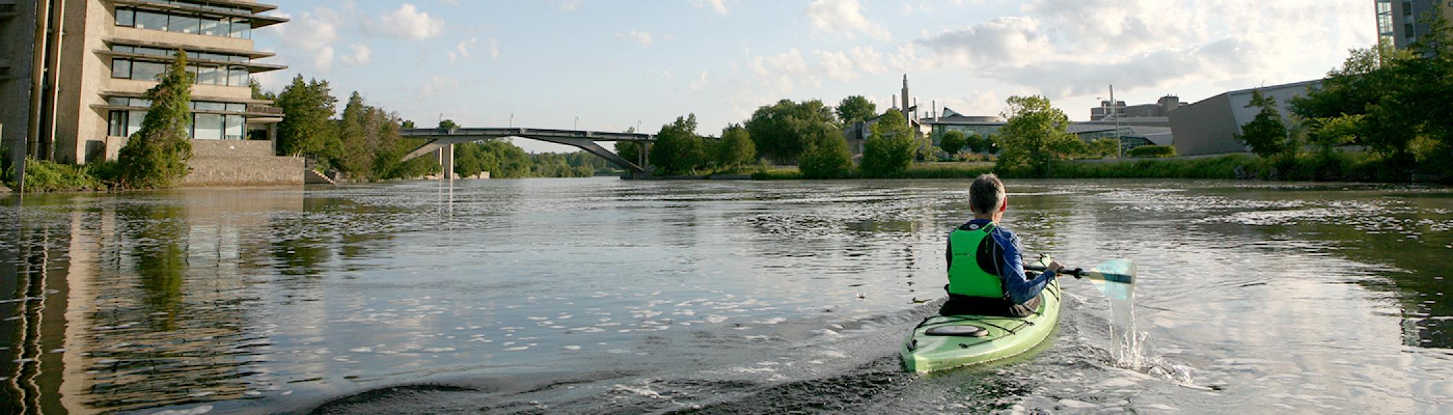 Dr. Leo Groarke kayaking on the Otonabee river in front of the Bata Library facing away from the camera towards the Faryon bridge