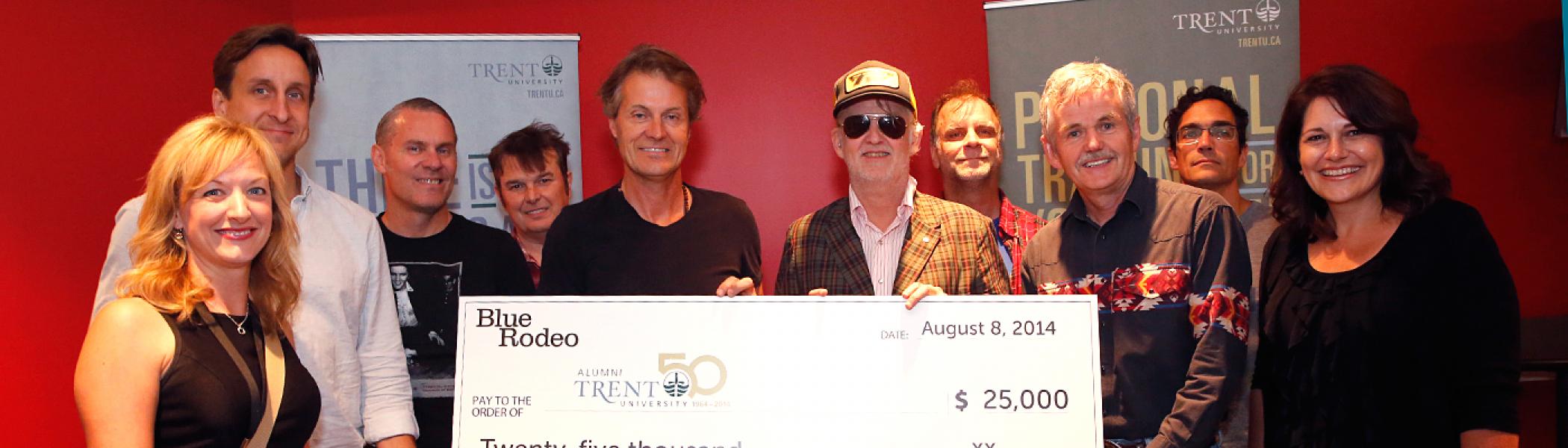 Dr. Leo Groarke, Julie Davis and Lee Hays posing with a large white check for $25,000 with the band Blue Rodeo in front of a red wall