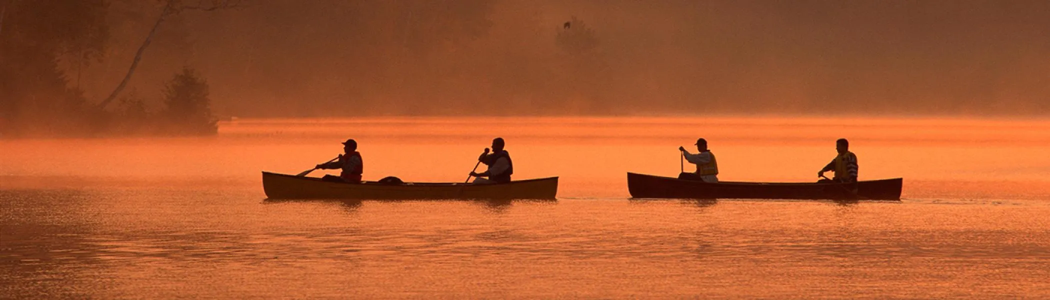 Two canoes with 2 people in each one, paddling acorss a lake at sunset
