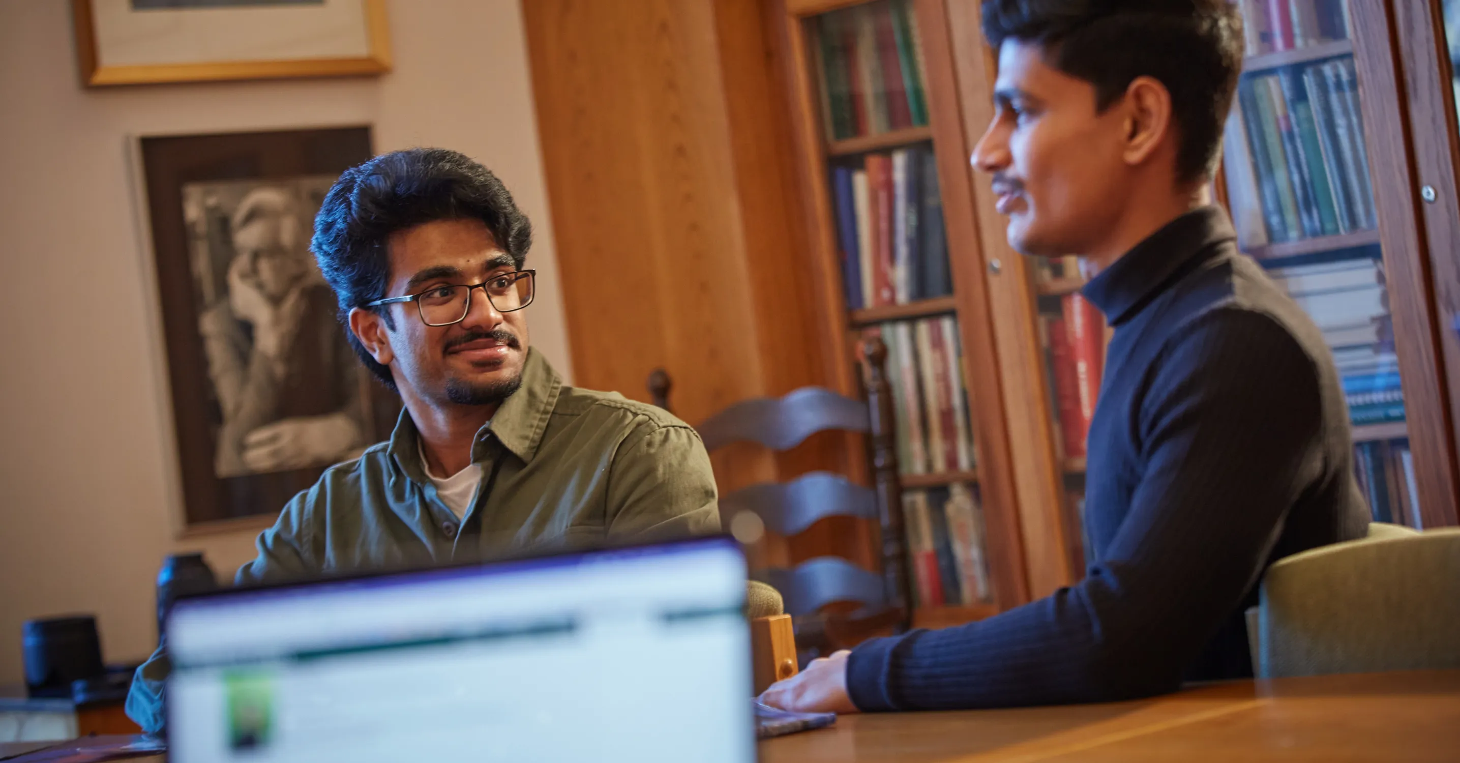 Two students sitting at a table in Kerr House at Traill College, talking to each other, with an out of focus computer screen in the foreground. 