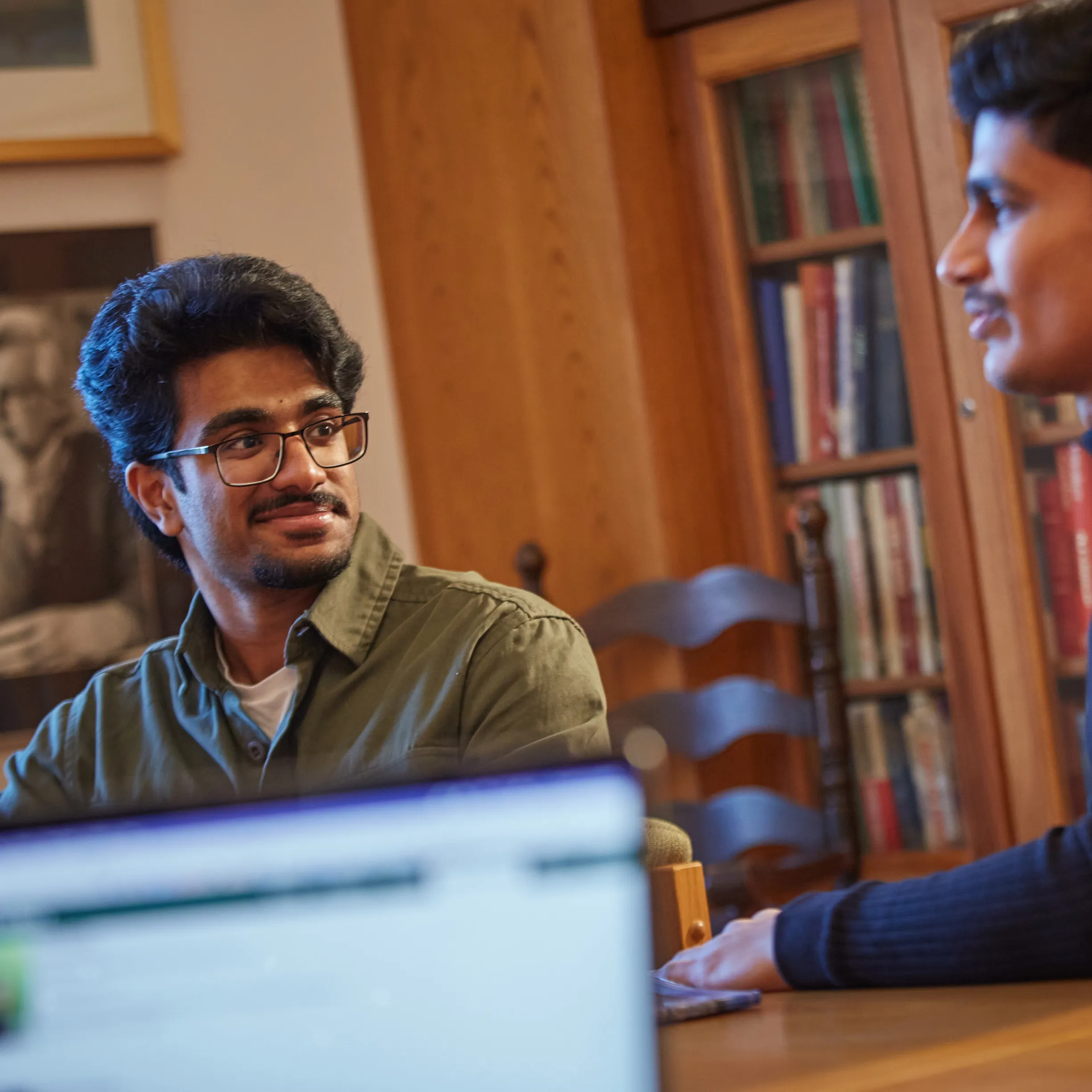 Two students sitting at a table in Kerr House at Traill College, talking to each other, with an out of focus computer screen in the foreground. 