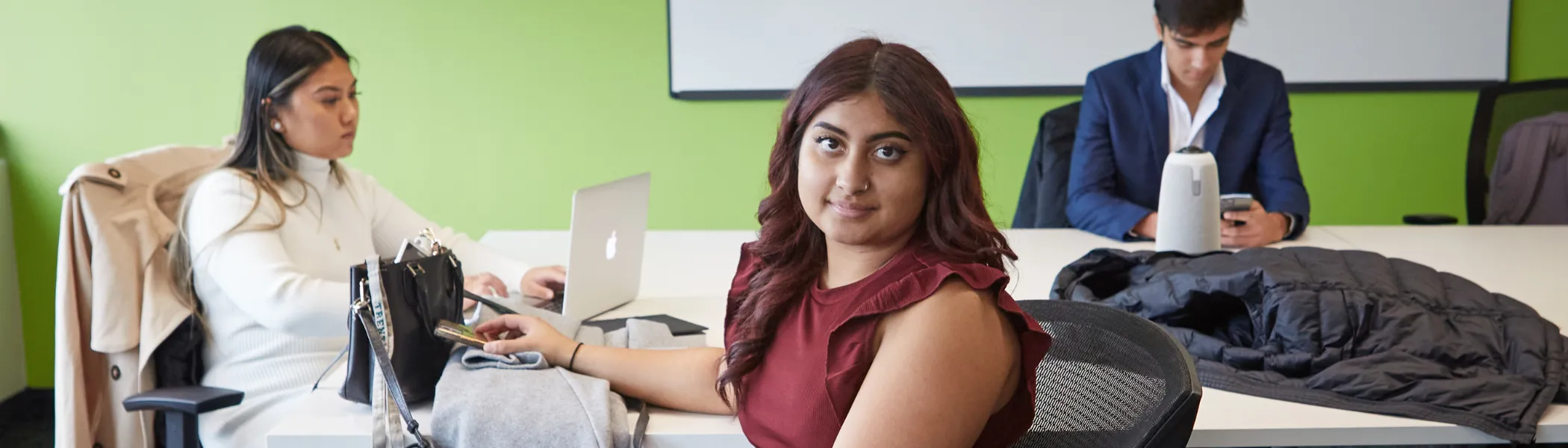 Three students sitting together at a table, in a office, with their laptops, student in the centre is looking at the camera