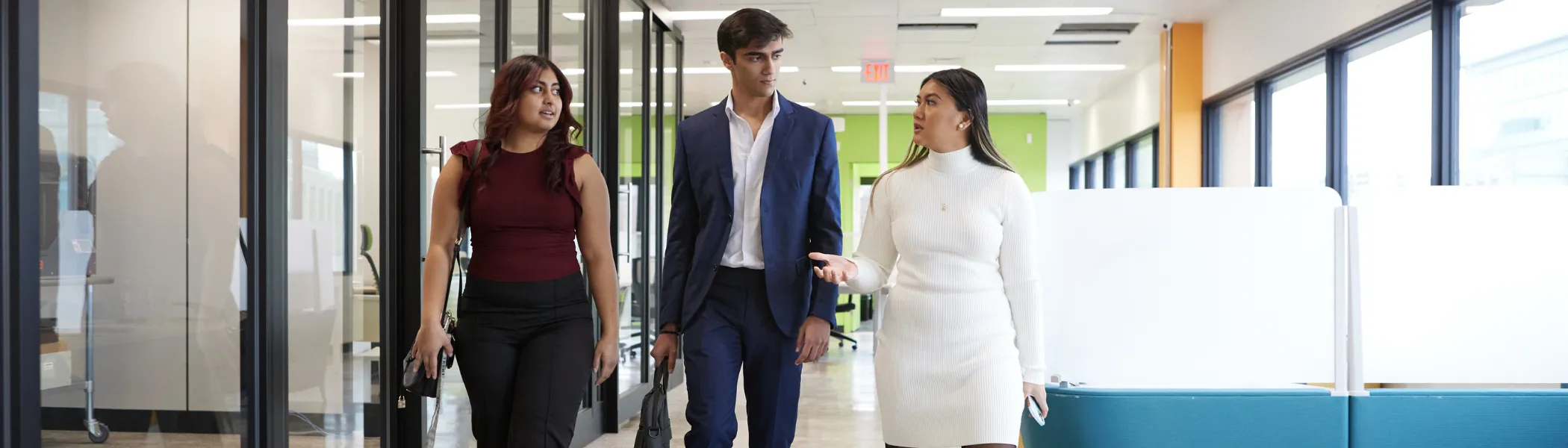 Three business students walking down an office hallway, talking to each other.