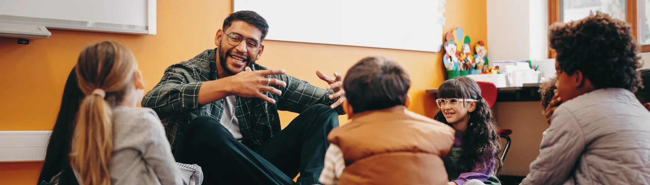 Image of a teacher sitting on the ground and talking with elementary school students.