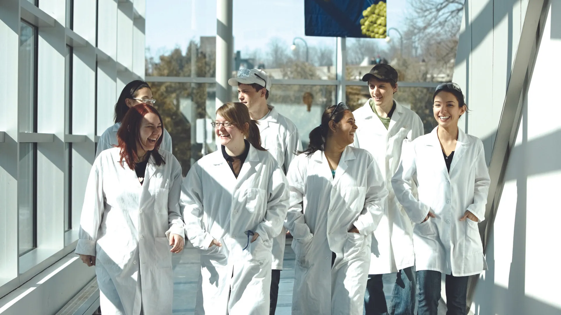 A group of Trent University students walking down the Chemical Science Building, wearing lab coats, and talking with each other. 