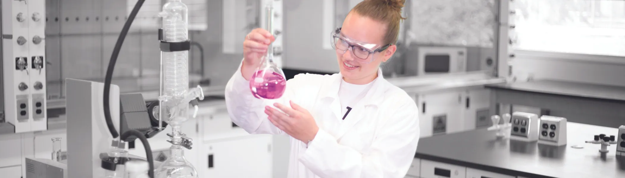 Trent University student in lab, holding up a chimerical glass beaker.