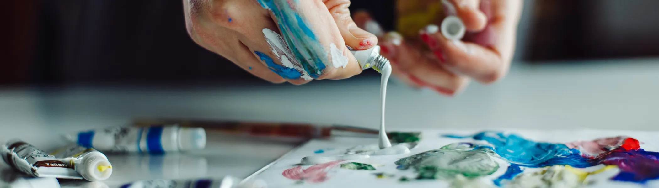 Hands cover in paint, pushing oil paint on palette on table.