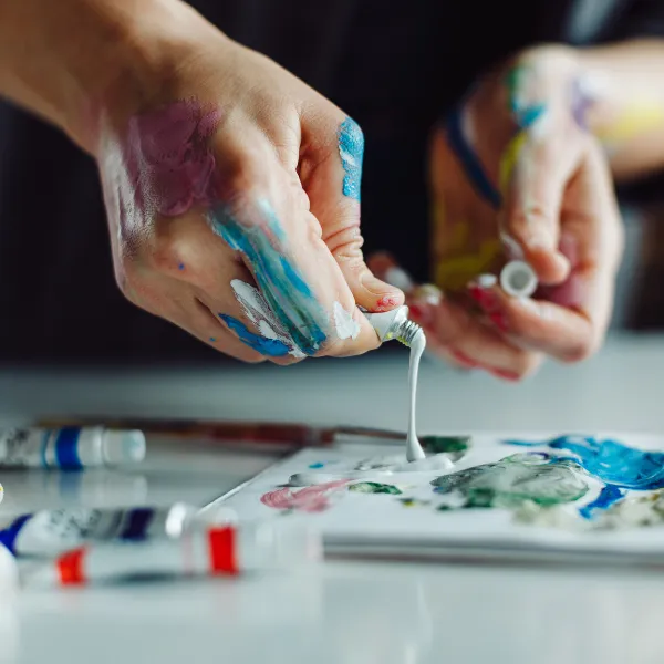 Hands cover in paint, pushing oil paint on palette on table.