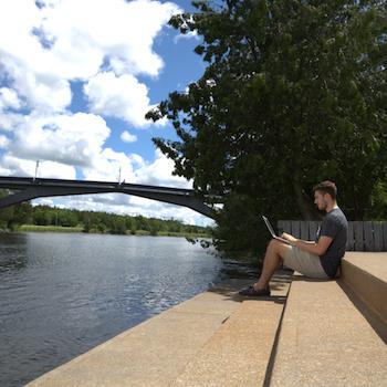 A guy sitting on steps by the river working on his laptop
