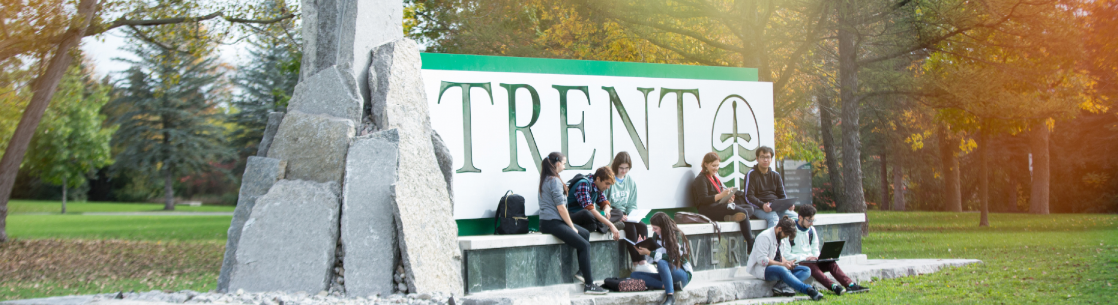 Main Trent University sign with students sitting nearby studying and laughing