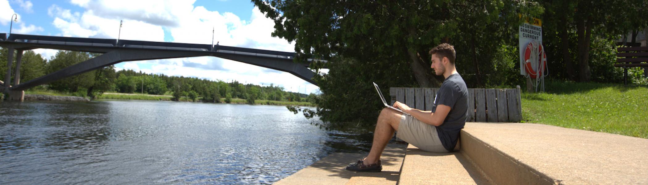 A guy sitting on steps by the river working on his laptop