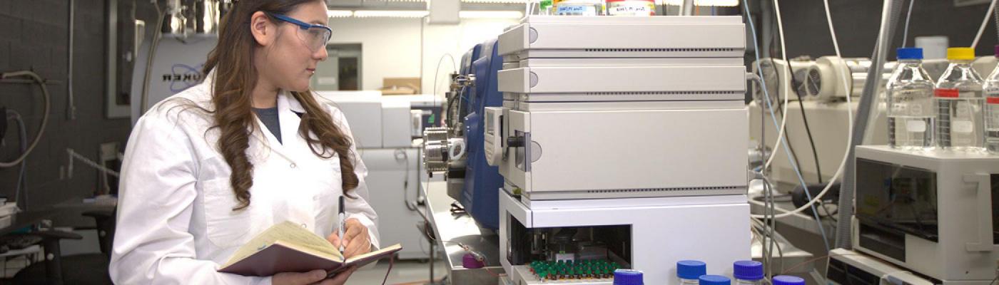 Scientist holding book and writing down results from equipment containing many vials 