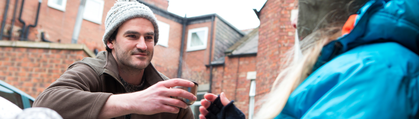 Photo of man kneeling down, extending cup to someone living on the street