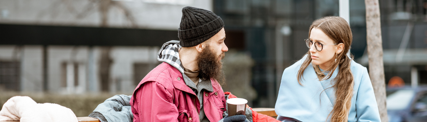 A homeless man sitting on a bench, with a coffee, talking to a younger woman who is listening intently