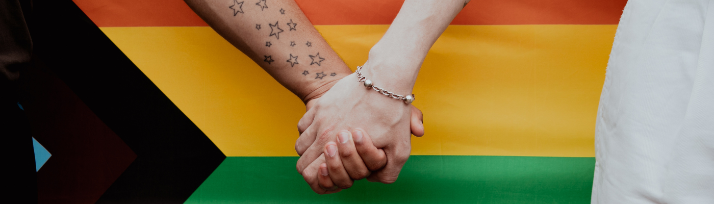 Close up image of two hands clasped together, in front of colours of pride flag
