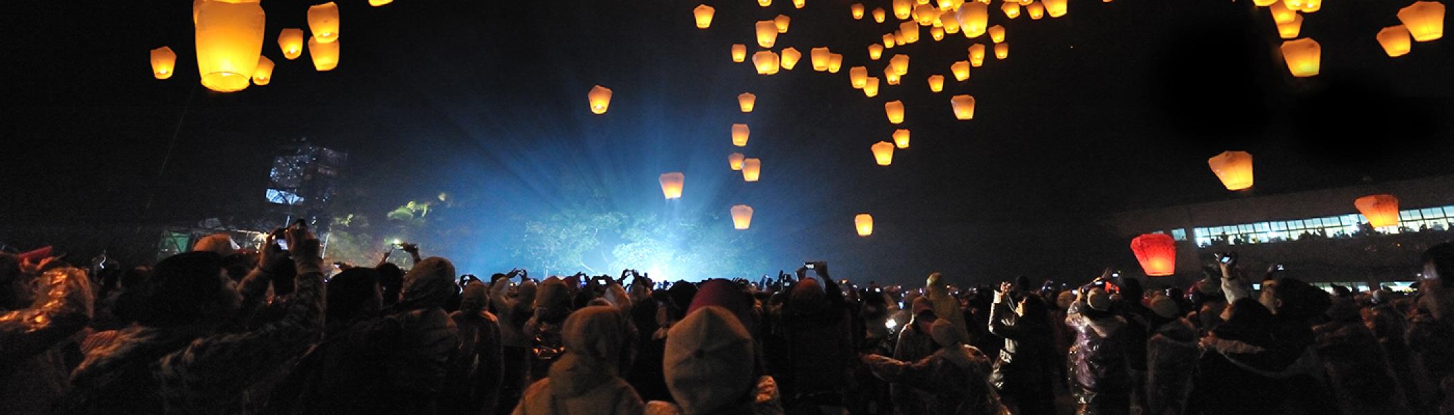 A crowd of people staring up a candles floating in the sky at night