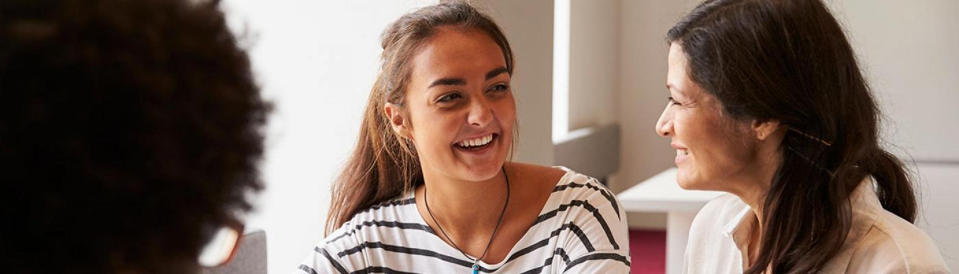 A student talking to a professor sitting beside her at a desk in front of a window